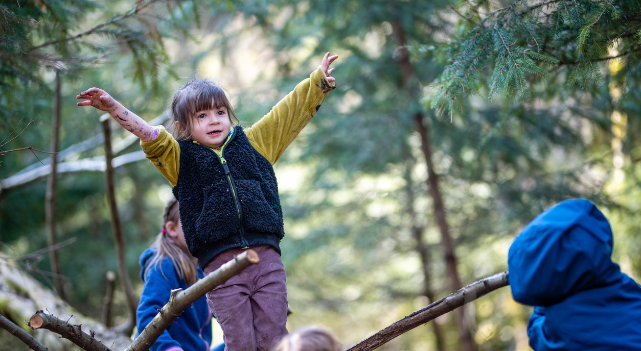 Ein Kind balanciert mit ausgestreckten Armen über einen umgefallenen Baumstamm im Wald